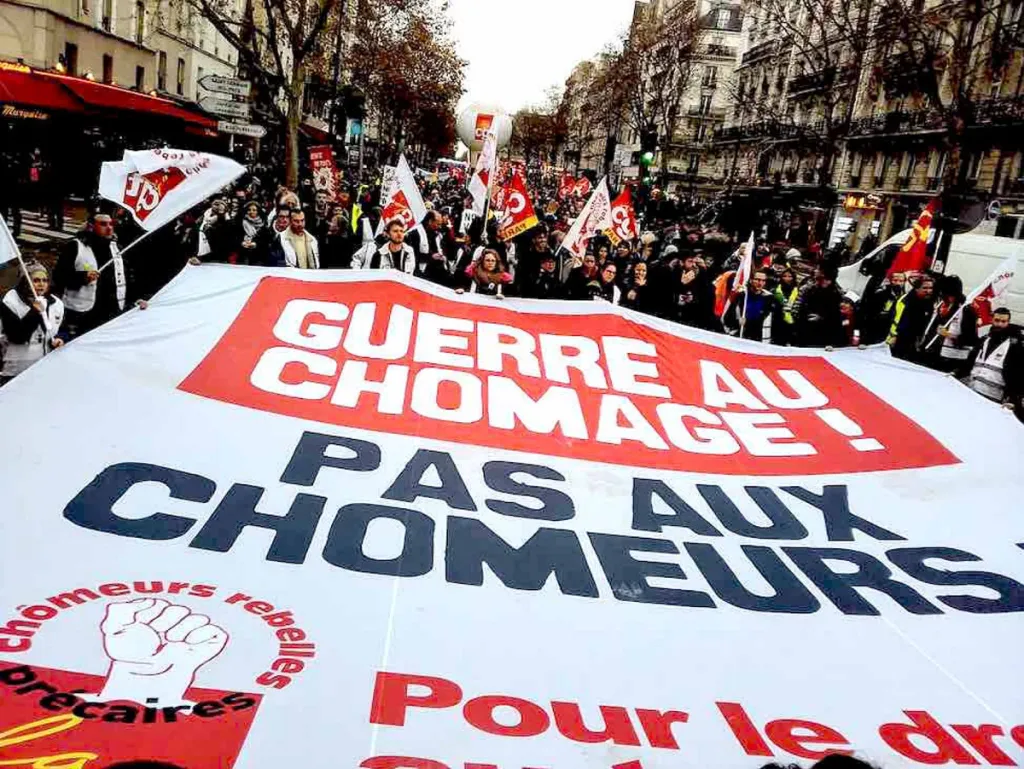 Photographie : cortège de manifestants derrière une grande banderole portant l'inscription « Guerre au chômage, pas aux chômeurs ! », drapeaux syndicaux à l'arrière-plan.