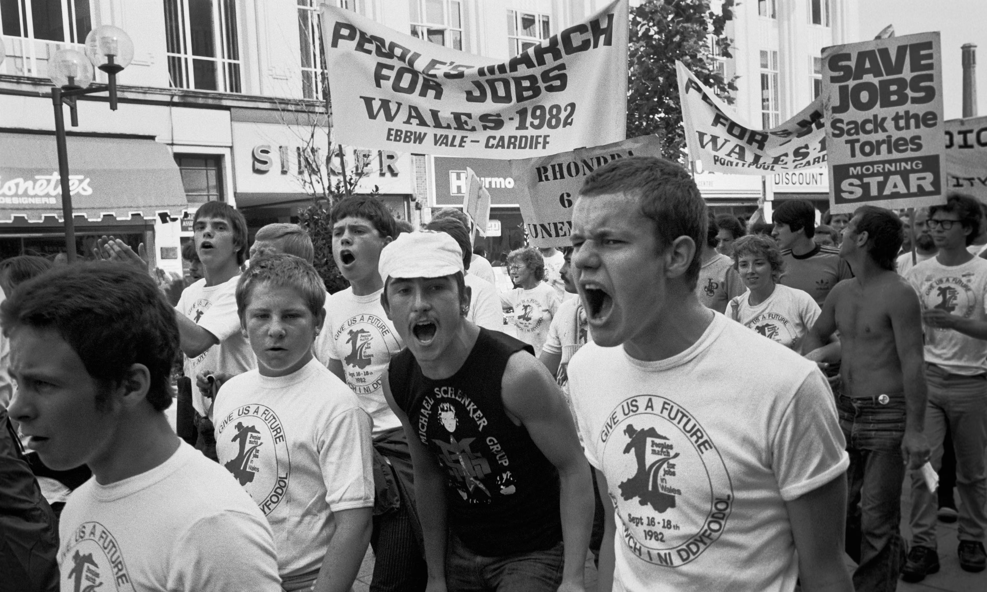 Photographie en noir et blanc : jeunes manifestants criant des slogans, portant des t-shirts « Give us a future » et des banderoles « People's March for Jobs, Wales 1982 ».