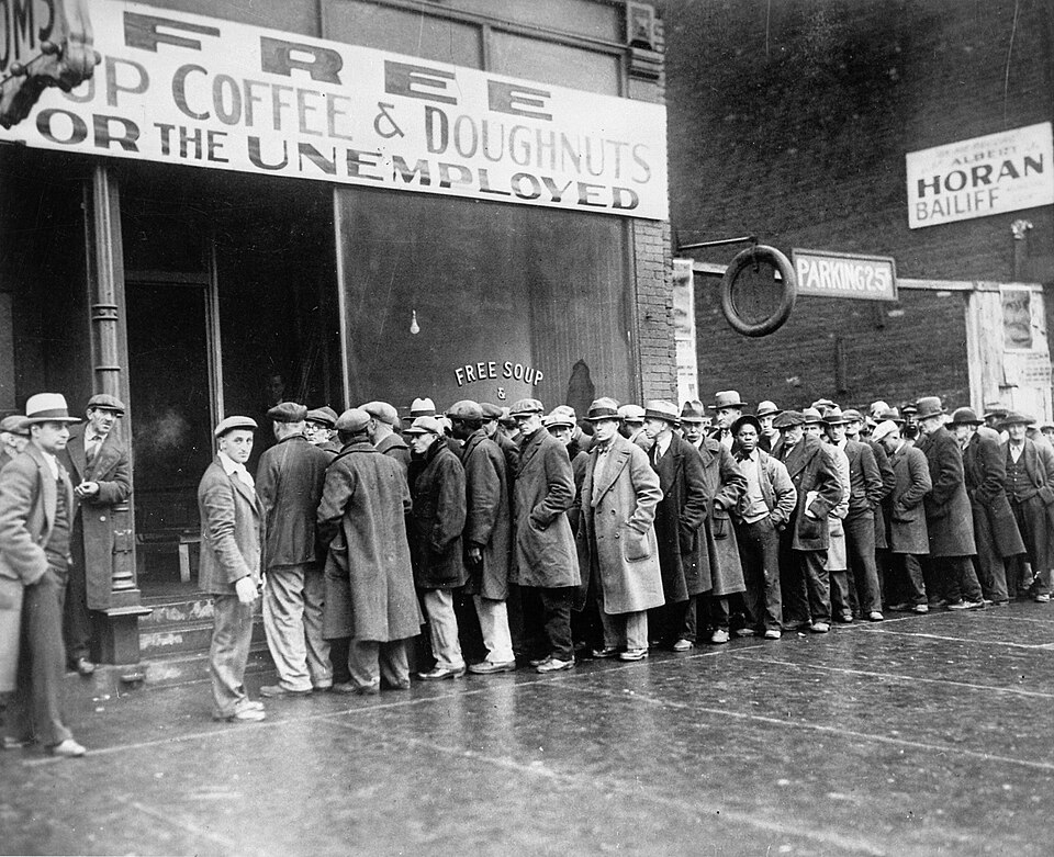 Photographie en noir et blanc : file d'hommes en manteau et chapeau attendant devant une devanture portant l'inscription « Free soup, coffee & doughnuts for the unemployed ».